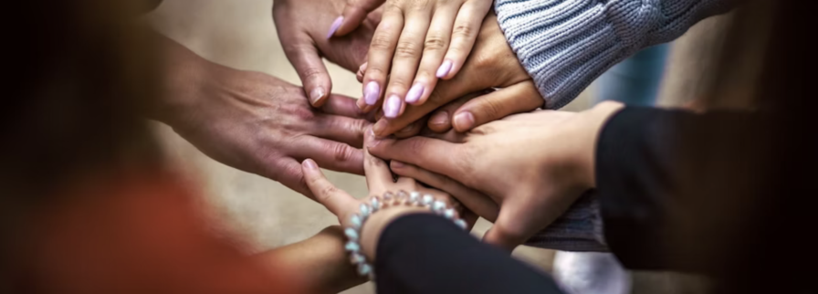 Een groep mensen staat in een cirkel en legt hun handen boven op elkaar in het midden, als teken van samenwerking en verbondenheid